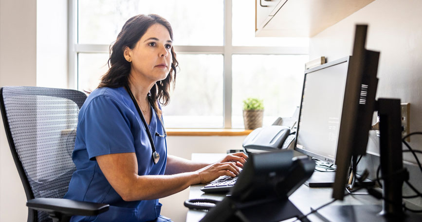 A healthcare professional sits at a desk and types on a keyboard while looking at a monitor screen.