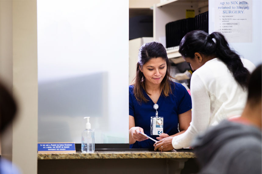 A female healthcare professional sits behind a counter in a provider waiting room and helps a woman with billing.