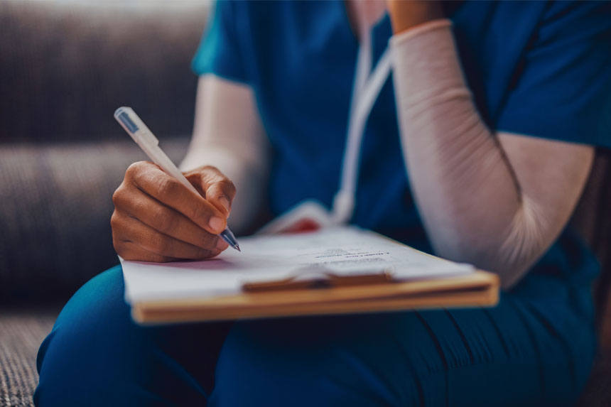 A close-up shot of the arms and torso of a healthcare worker in scrubs and a long undershirt, holding a pen and filling out a form on a clipboard.