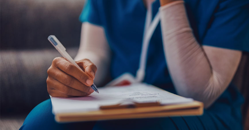 A close-up shot of the arms and torso of a healthcare worker in scrubs and a long undershirt, holding a pen and filling out a form on a clipboard.