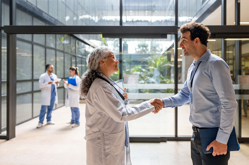 A female doctor and male medical science liaison shake hands in the well-lit lobby of a hospital.