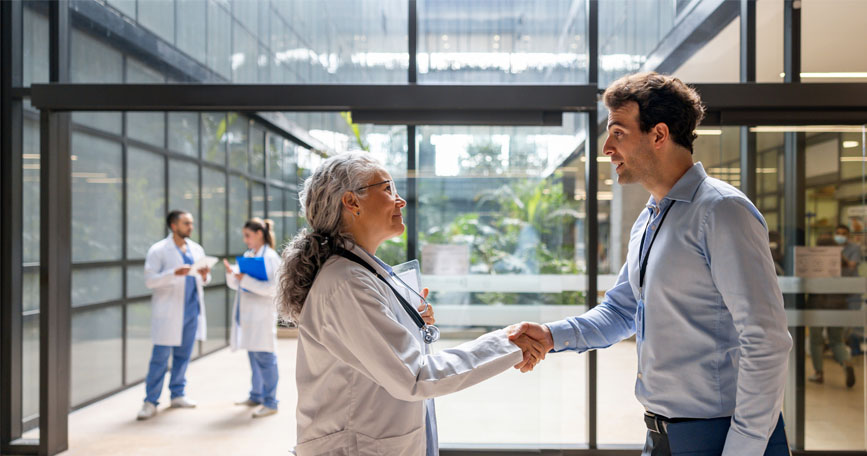 A female doctor and male medical science liaison shake hands in the well-lit lobby of a hospital.