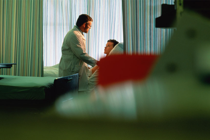 A doctor stands at a patient's bedside and provides care in a dimly lit hospital room.