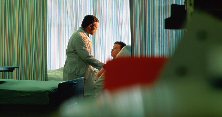 A doctor stands at a patient's bedside and provides care in a dimly lit hospital room.