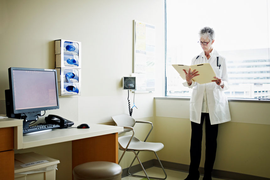 A female clinician stands near the window of a well-lit exam room and looks over a medical chart.