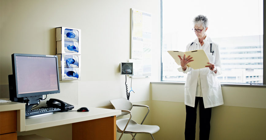 A female clinician stands near the window of a well-lit exam room and looks over a medical chart.