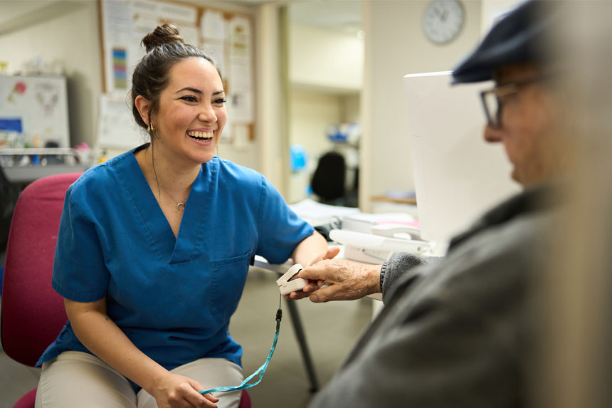 A female healthcare worker in blue scrubs smiles as she takes the pulse oximetry reading of an elderly male patient.