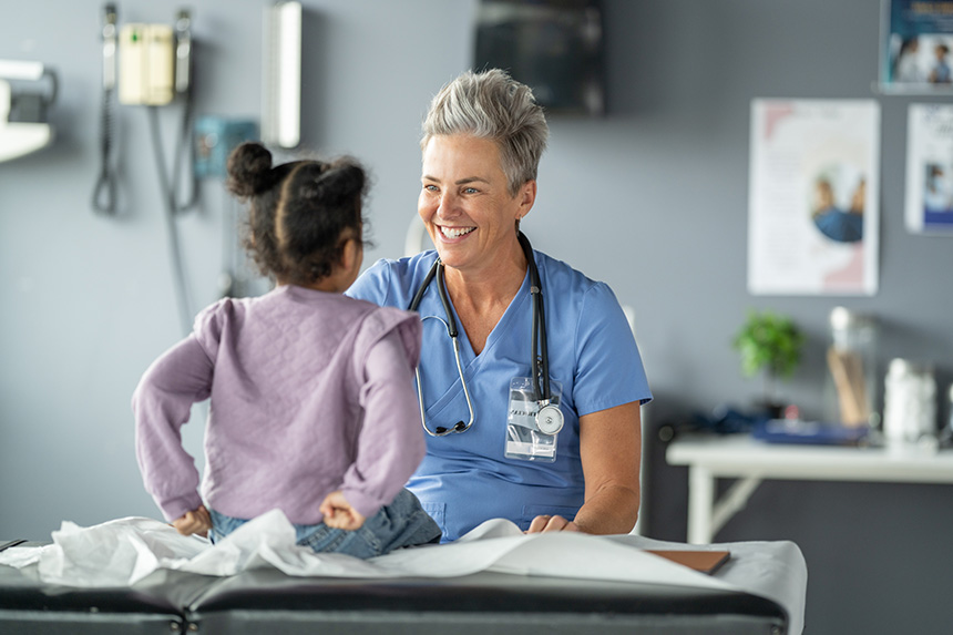 A female clinician with short grey hair sits in front of a young child seated on an exam table and smiles.