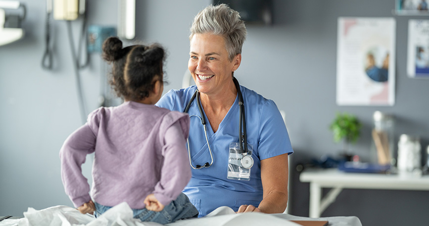 A female clinician with short grey hair sits in front of a young child seated on an exam table and smiles.