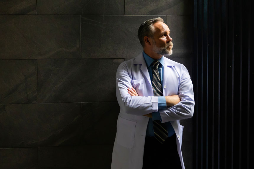 A male healthcare professional in a white coat looks up and to the right while standing in a dark hallway.