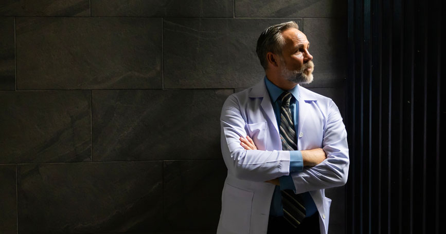 A male healthcare professional in a white coat looks up and to the right while standing in a dark hallway.