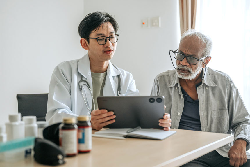 A younger male physician sits at a desk beside an older male patient, using a tablet to provide digital health care.