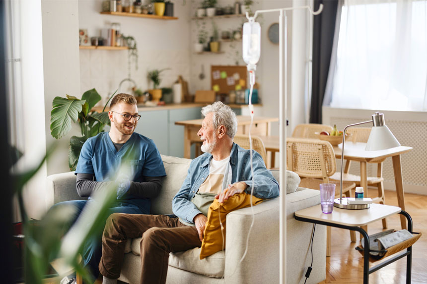 A home healthcare worker and his elderly patient sit on a couch together in a well-lit living room. 