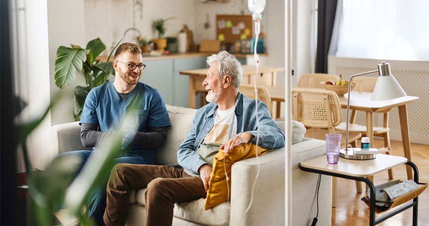 A home healthcare worker and his elderly patient sit on a couch together in a well-lit living room. 