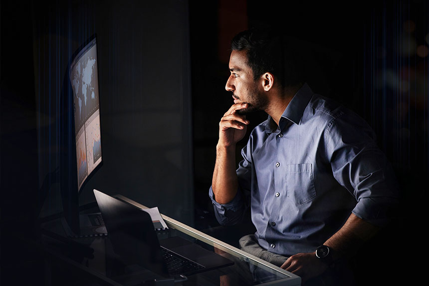 A male dressed in business attire sits in a dark room in front of a lit screen.