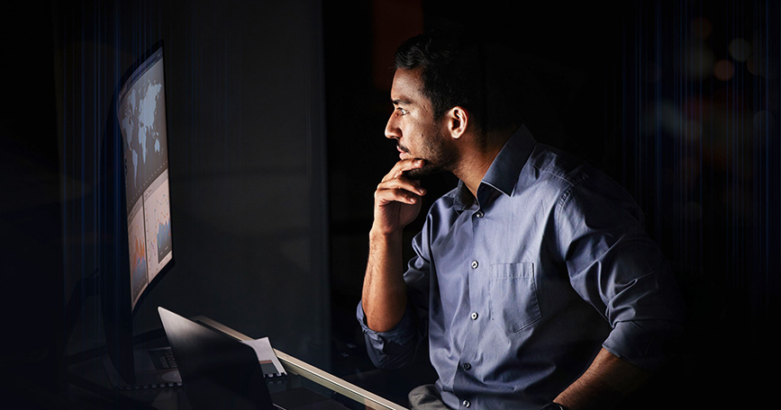 A male dressed in business attire sits in a dark room in front of a lit screen.