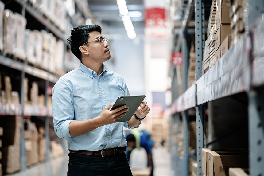 A worker examines a shelf in a warehouse and takes inventory on a tablet. 