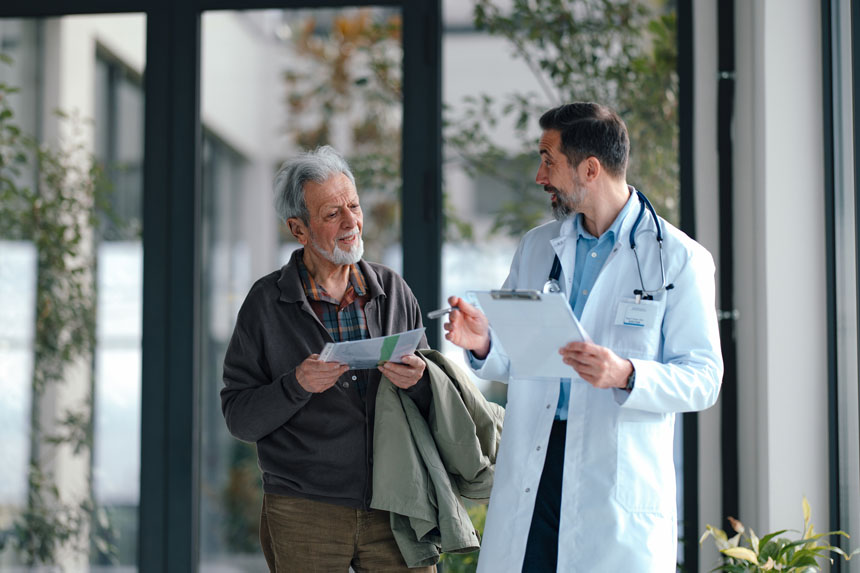 An elderly patient in casual clothing chats with a doctor in a white coat about his charts.