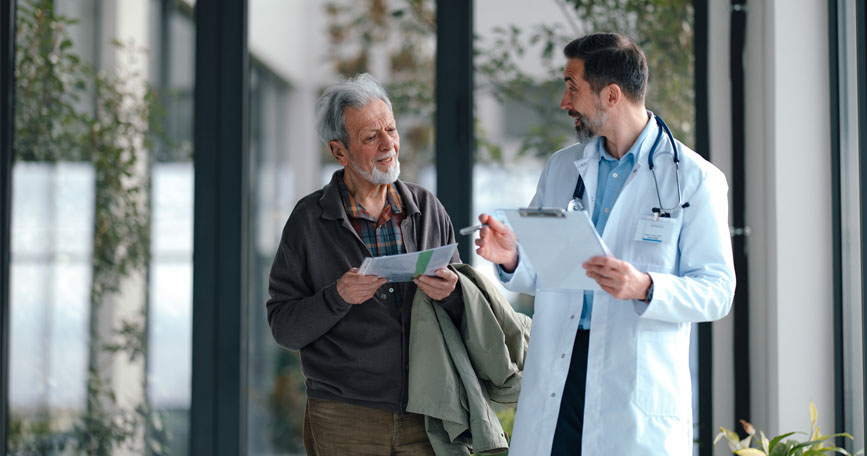 An elderly patient in casual clothing chats with a doctor in a white coat about his charts.