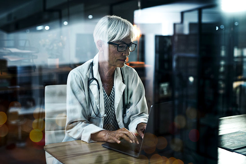 A female healthcare professional in a white lab coat sits at a table while typing on a laptop keyboard.