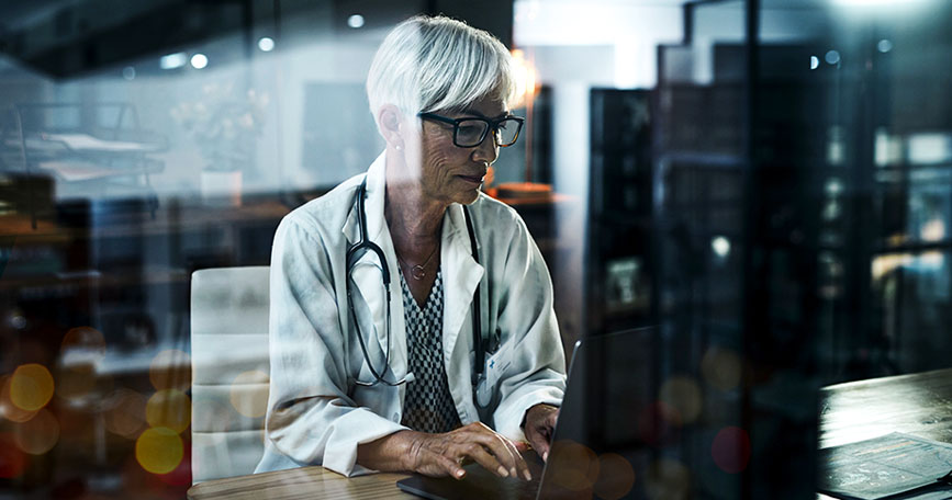 A female healthcare professional in a lab coat sits at a table while typing on a laptop keyboard.