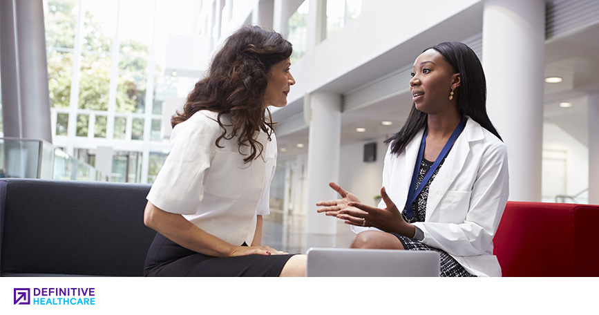 A white woman in a blouse talking to a black woman in a white doctor's coat