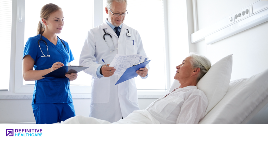 Two clinicians stand at a patient's bedside and look at his charts. 