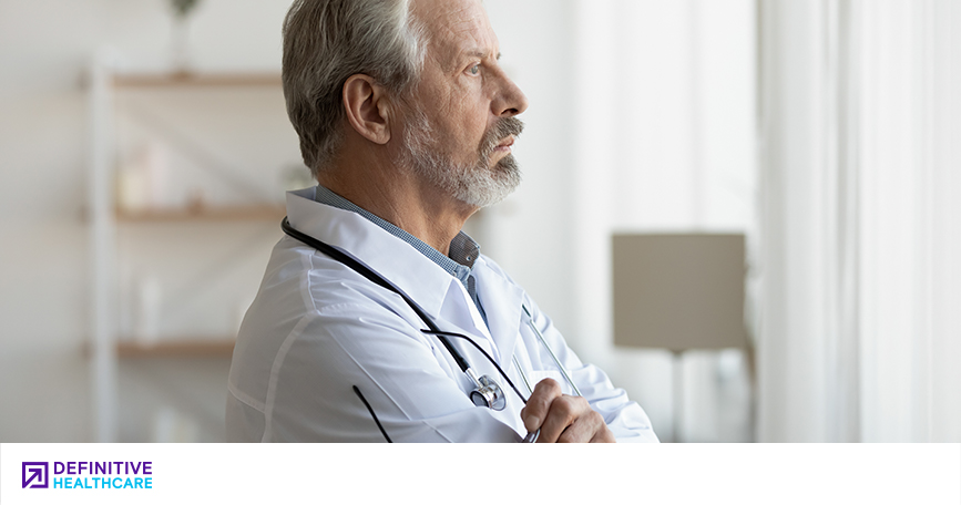 An older white male doctor looking out a window with a thoughtful expression