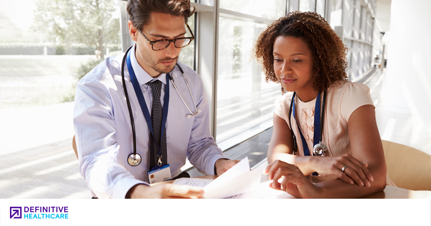 A white man in glasses with in a white dress shirt and tie with stethoscope around his neck sits to a Black woman in a short sleeve shirt and a stethoscope around her neck are sitting at a table looking at papers