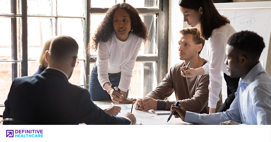 A diverse group of men and woman standing and sitting around a table in a work meeting