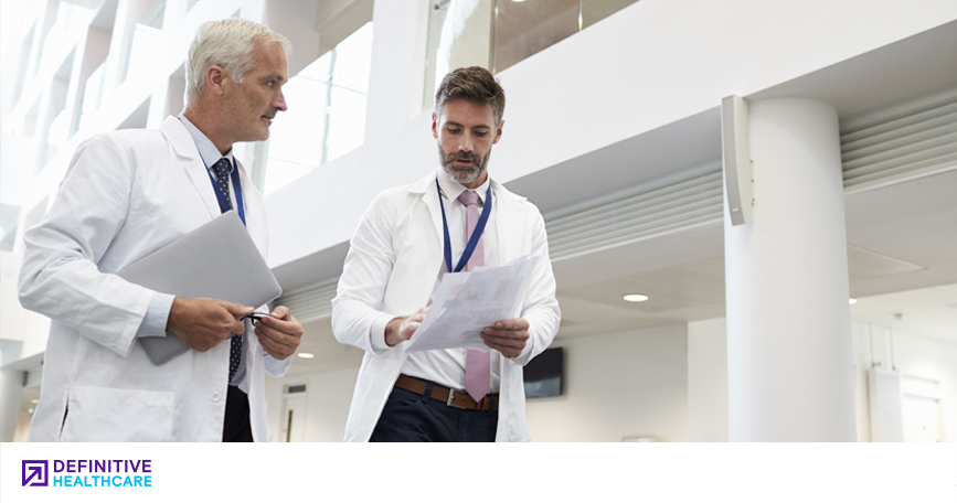 A white man with white and a white man with dark hair wearing white lab coats walk in a white hallway with papers in their hands