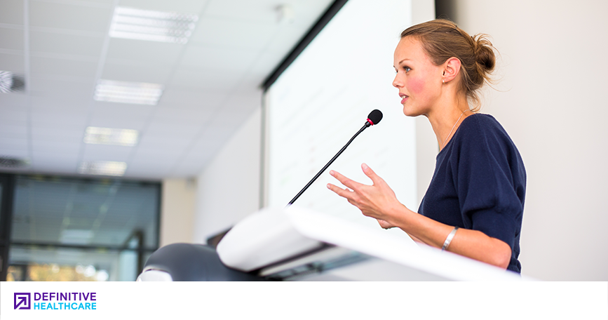 A woman speaks into a microphone at a lectern.