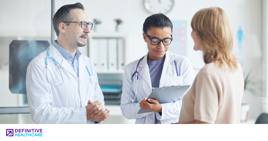 A clinician speaks to a patient with his hands clasped while another clinician writes on a clipboard.