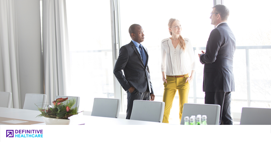 Three people wearing business attire speak together in a meeting room.