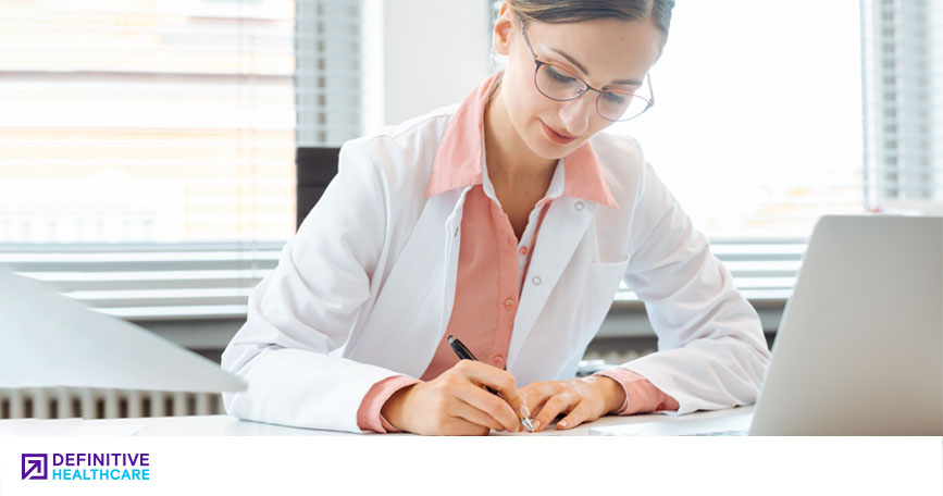A female healthcare professional sits at a desk and takes notes with a pen. 