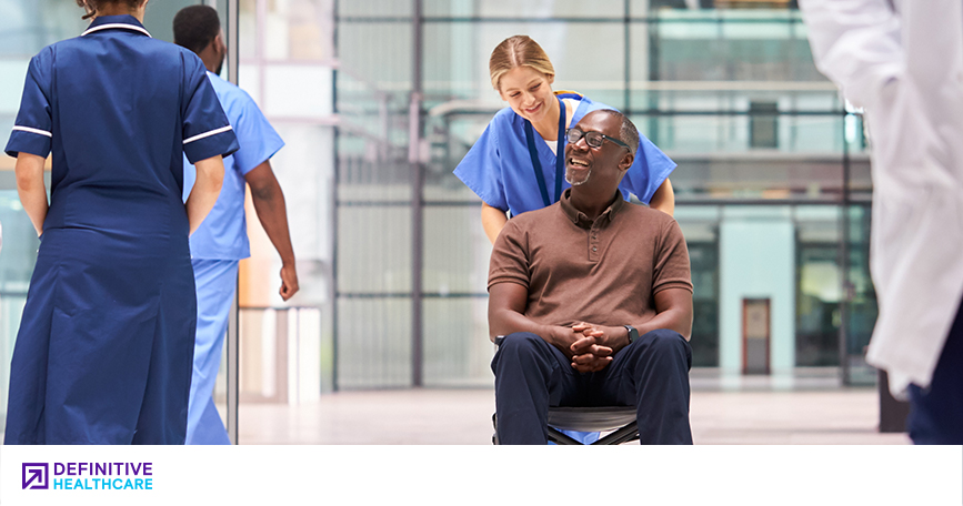 A woman in scrubs pushing a man in a wheel chair out of a healthcare facility