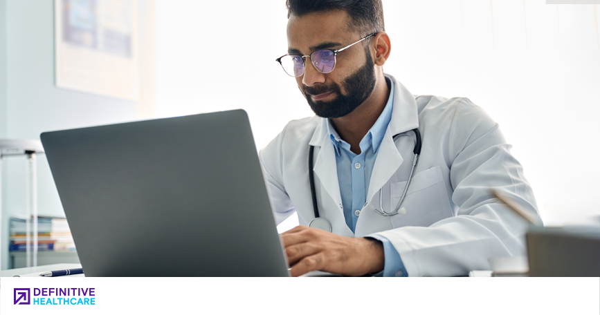 A male doctor in a white coat with a stethoscope around his neck sits and looks at a computer