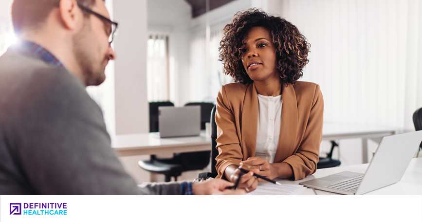 A woman in business attire speaks to a man in a professional setting.