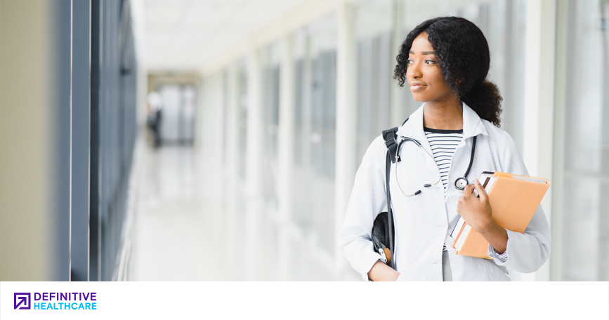 A young woman wearing a lab coat and stethoscope walks down a hallway. 