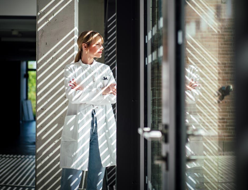 A healthcare provider stands, looking out a window with her arms folded.