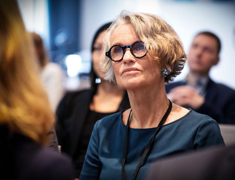 A woman with black frame round glasses sits and looks ahead. There are other people sitting near her.