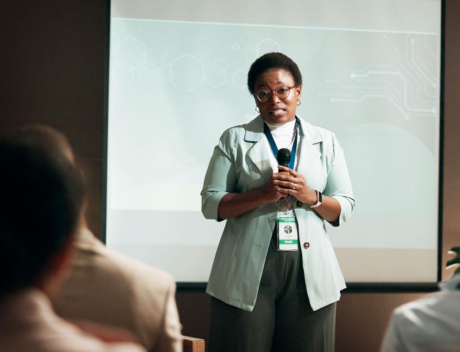 A woman in business casual attire stands in front of a projector screen holding a microphone while looking out at an audience.