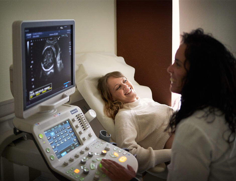 A female patient lays on an exam room table smiling as a technician performs an ultrasound.