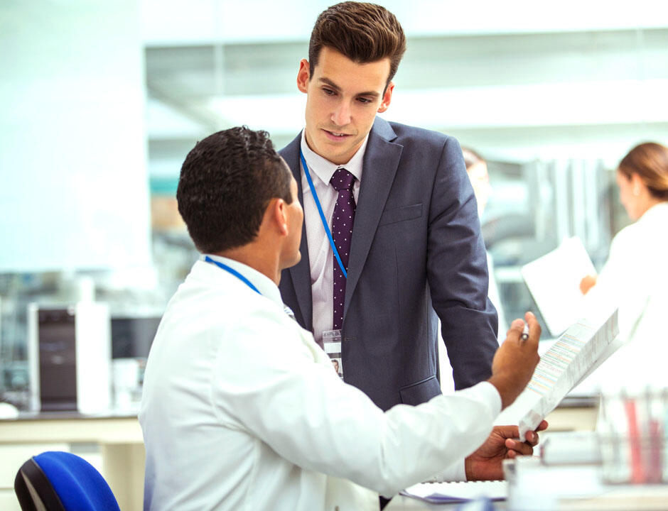 A male dressed in business attire stands at a table while talking to a healthcare professional in a white coat.