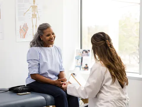 A female patient sits on an exam room table smiling as her physician sits in front of her holding a clipboard with papers.