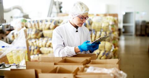 A person with glasses in a white coat, hair net and blue gloves is holing a table surrounded by boxes