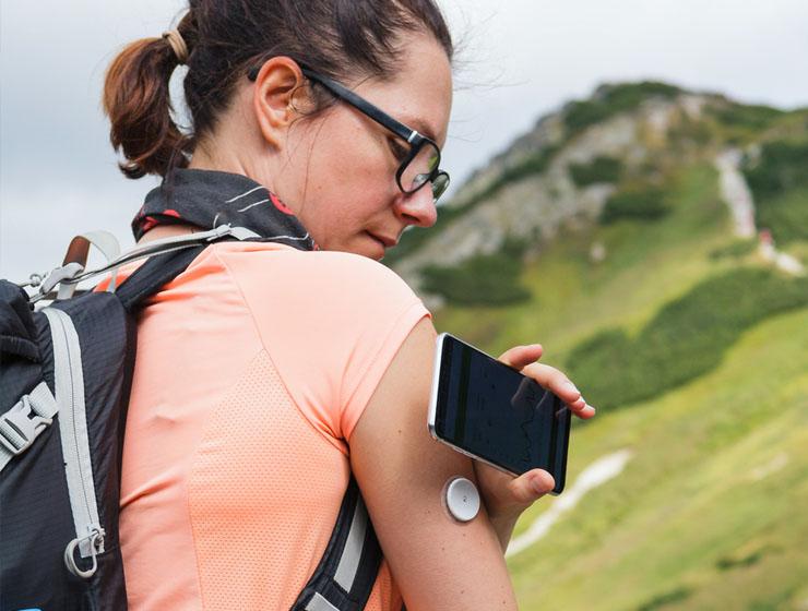 A woman with a backpack stands near a mountain as she places her phone near a glucose monitor on her arm.