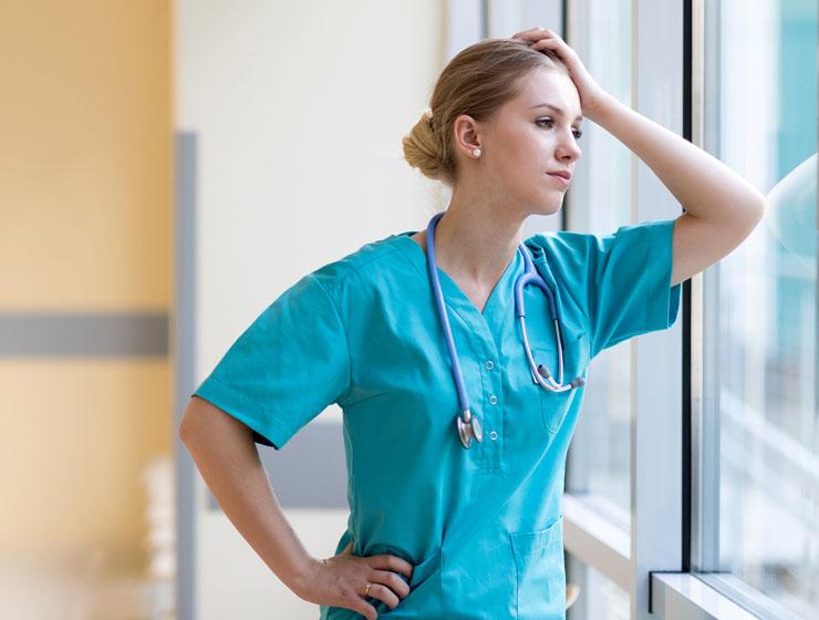 A female healthcare professional stands at a window with her elbow placed on the glass and hand touching her head.