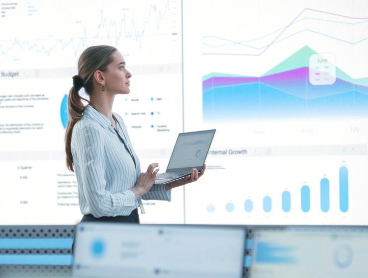 A woman in business attire stands with a laptop in her hands as she looks at a presentation of charts and graphs on a screen.