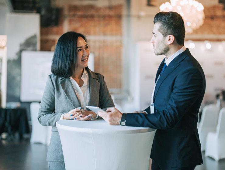 A man and woman in business attire stand at a table facing each other. The man is holding a tablet in one hand.
